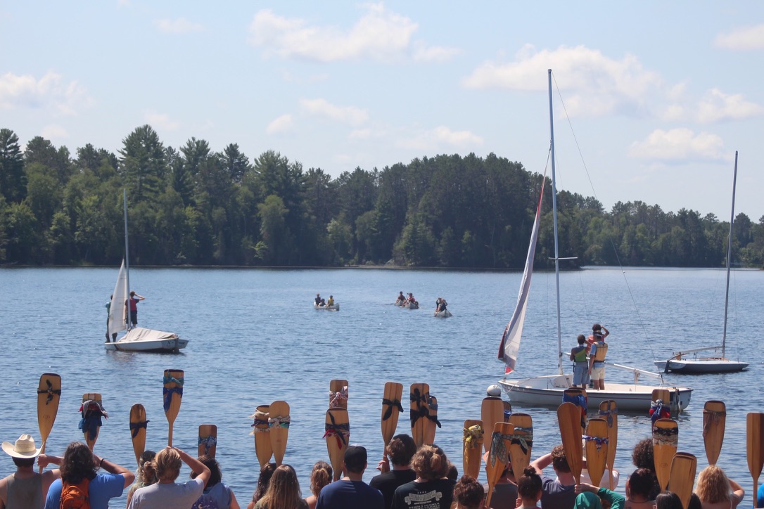 Campers return from 10-day trip to the Boundary Waters. Counselors who have taken the trip hold up oars with bandanas to salute the returning campers. Each bandana is unique, representing a completed trip.